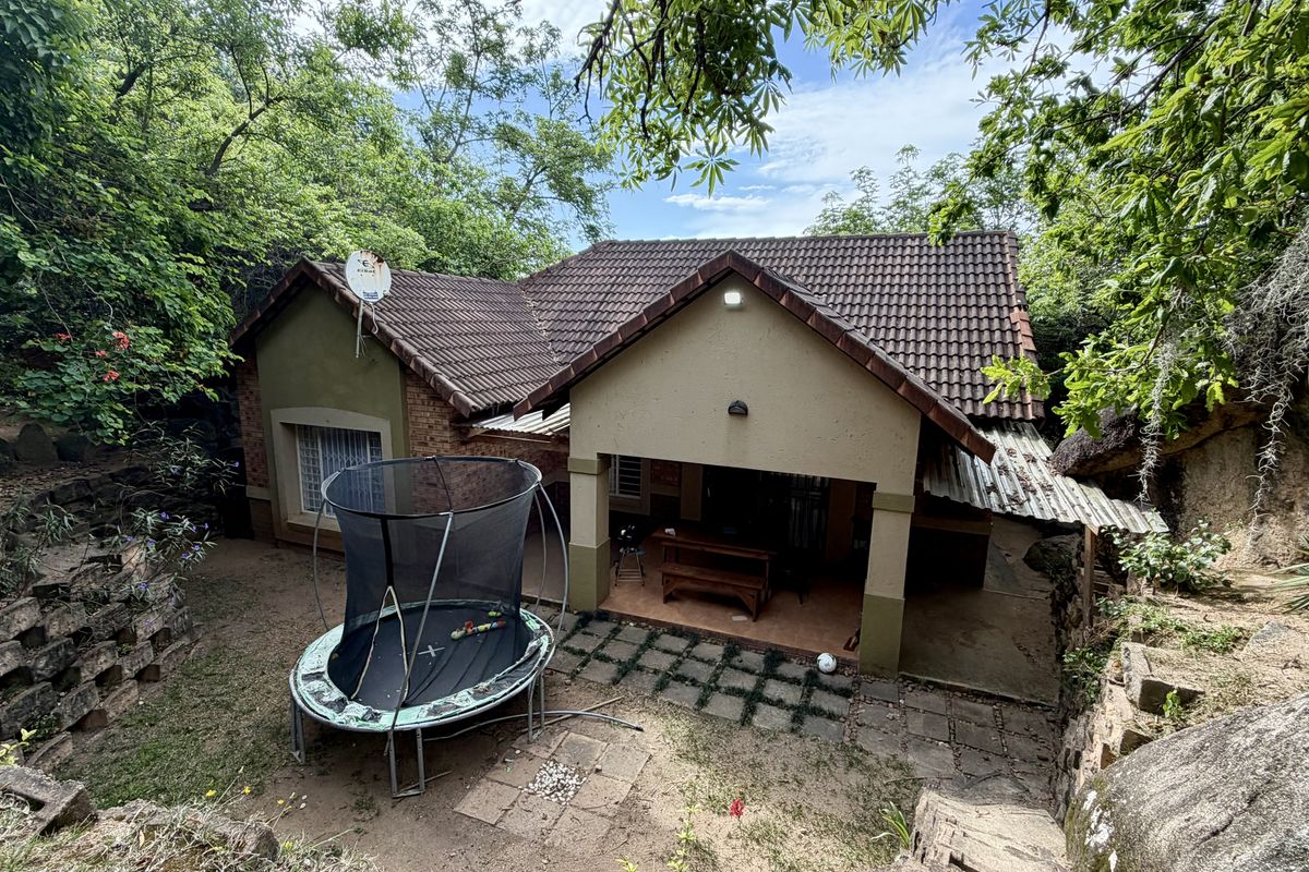 Rear view of a single-story house featuring a tiled roof, covered patio area, and natural garden surroundings with rock formations.