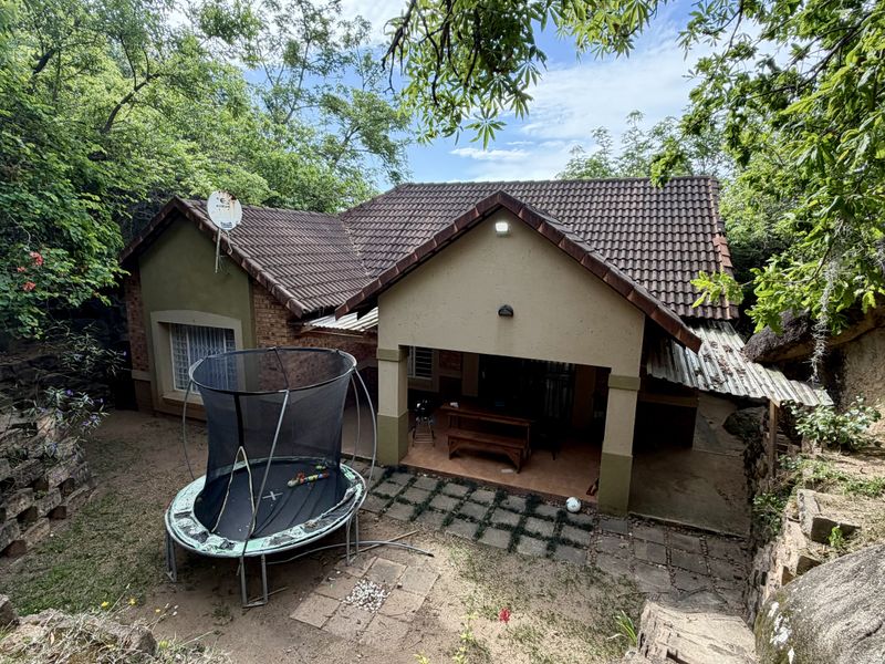 Rear view of a single-story house featuring a tiled roof, covered patio area, and natural garden surroundings with rock formations.