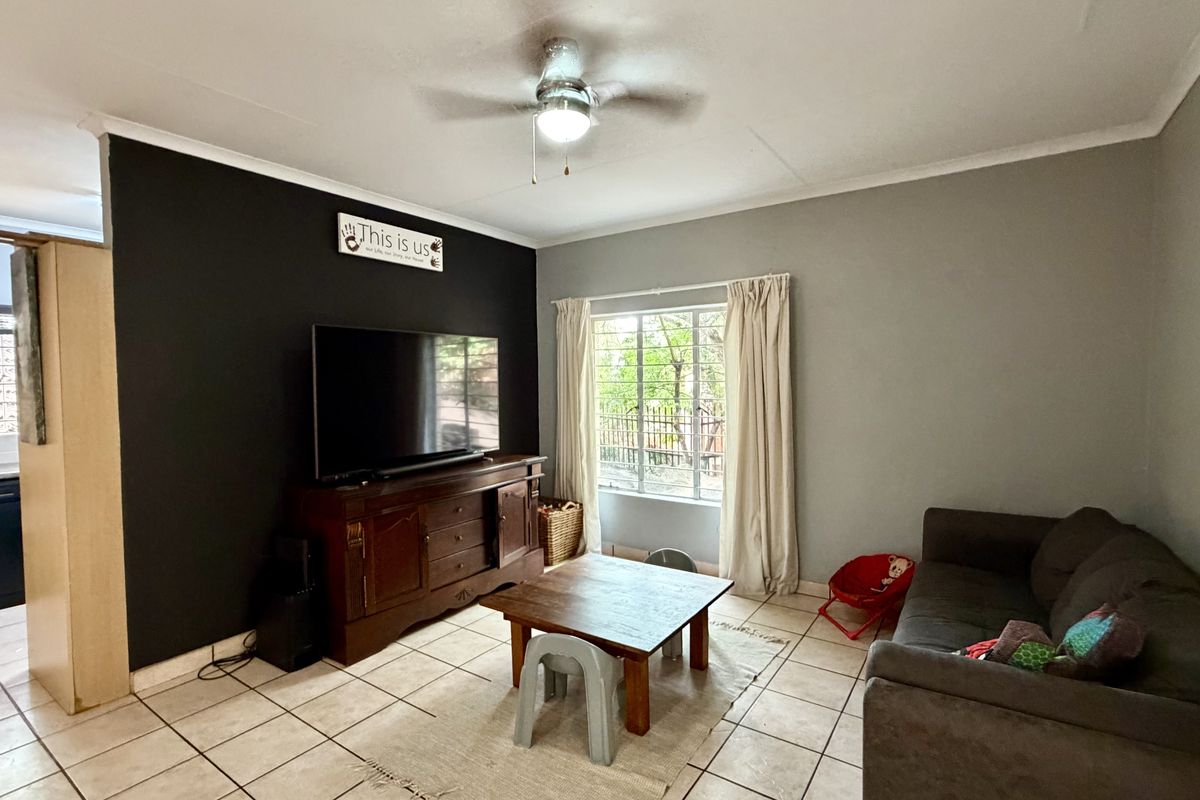 A living room featuring tiled flooring, a ceiling fan, and a window with security bars, adjacent to a kitchen area.