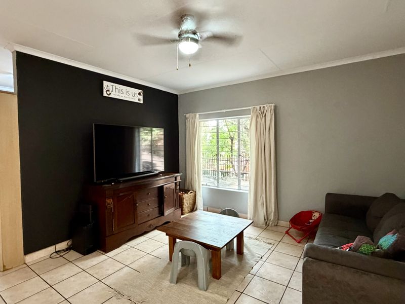 A living room featuring tiled flooring, a ceiling fan, and a window with security bars, adjacent to a kitchen area.