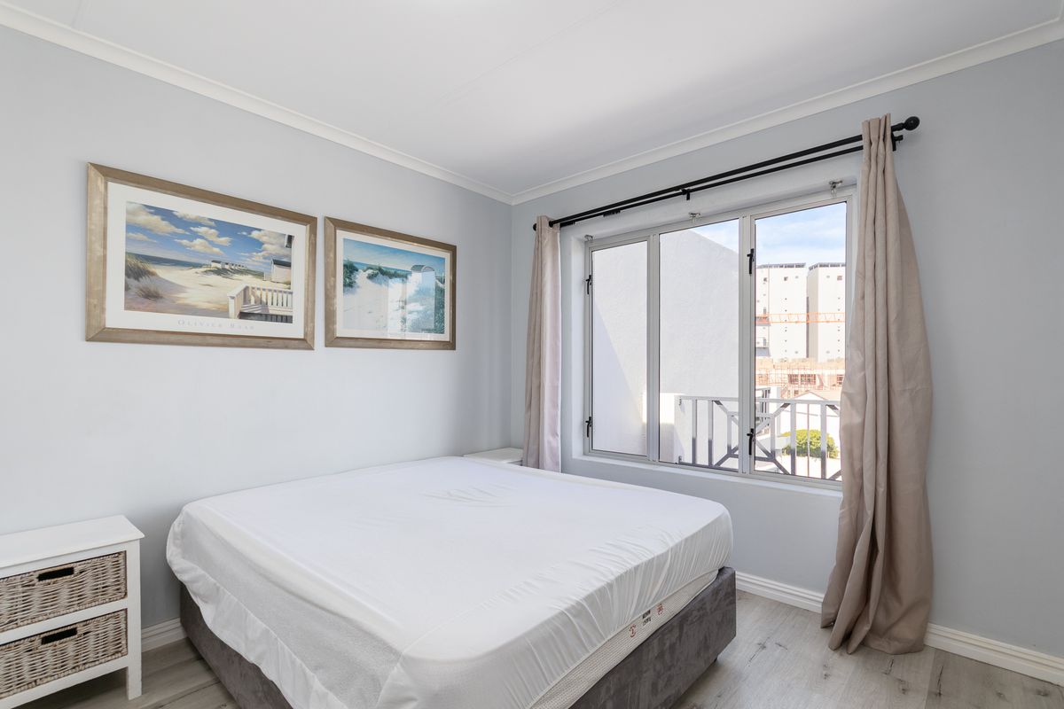 A bright and well-maintained bedroom featuring light-toned laminate flooring, neutral wall finishes, and a large aluminium-framed window allowing for natural light.