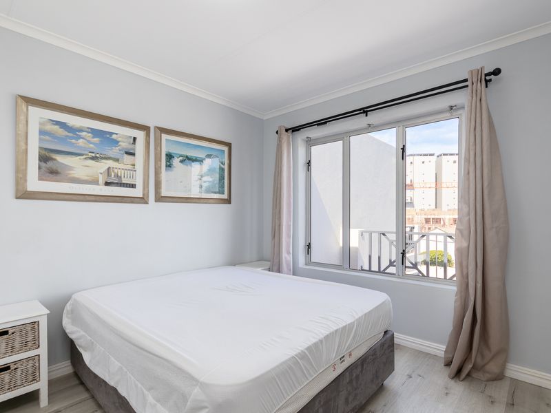 A bright and well-maintained bedroom featuring light-toned laminate flooring, neutral wall finishes, and a large aluminium-framed window allowing for natural light.