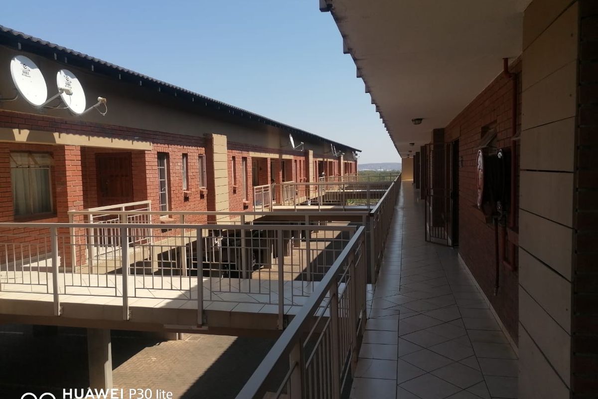 An exterior view of an apartment complex walkway featuring brick facade construction, tiled flooring, and metal safety railings.