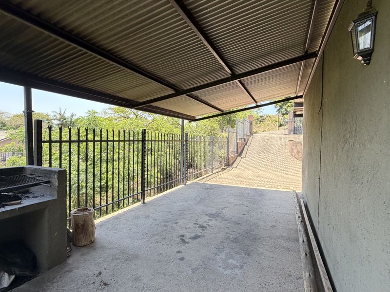 A covered outdoor patio area featuring a built-in braai, concrete flooring, and metal roofing, overlooking a paved driveway and surrounding greenery.