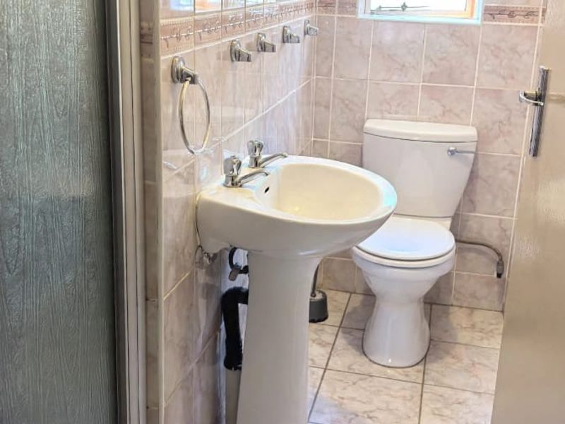 A functional bathroom featuring a pedestal basin, toilet, and shower enclosure with neutral ceramic wall and floor tiling.