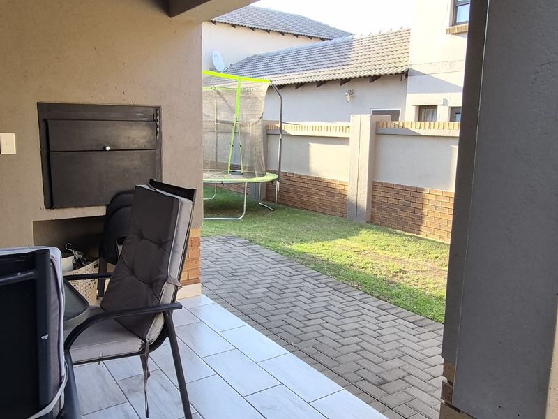 A covered patio featuring tiled flooring and a built-in braai, overlooking a paved walkway and a private garden area.