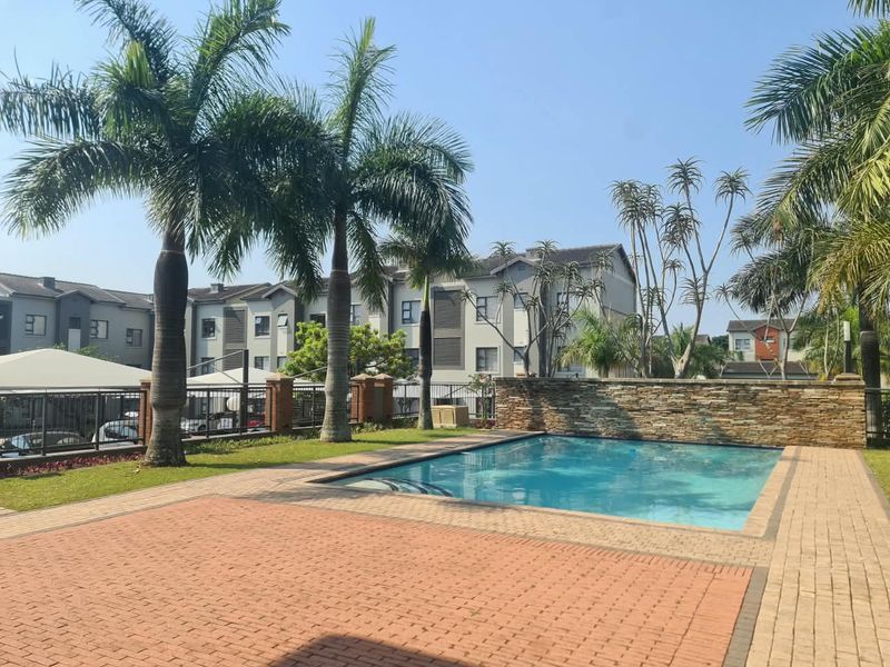 A well-maintained communal swimming pool area featuring a stone accent wall, brick paving, and established palm trees within an apartment complex setting.