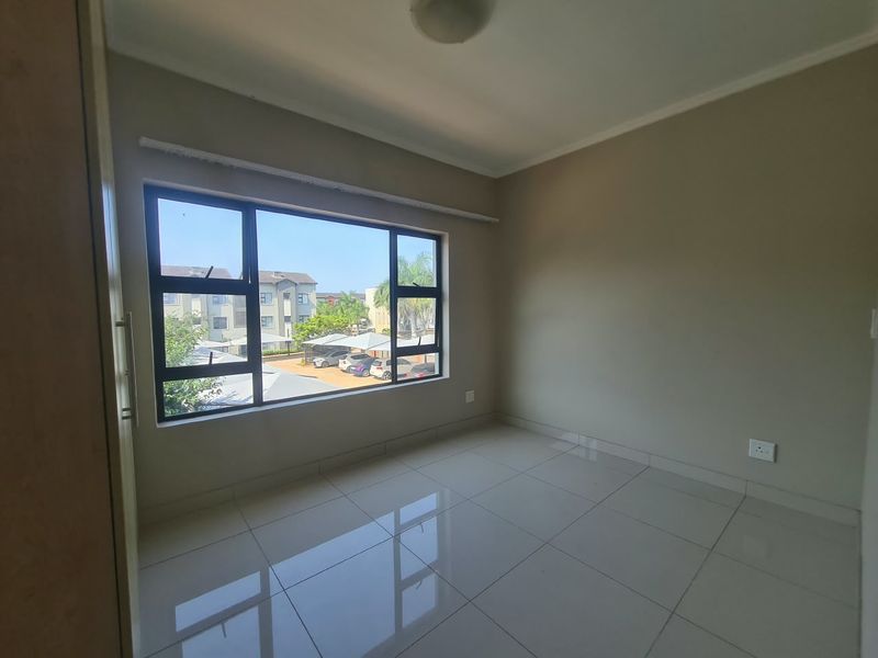 An empty bedroom featuring tiled flooring, neutral wall paint, and large aluminium-framed windows overlooking the complex parking area.
