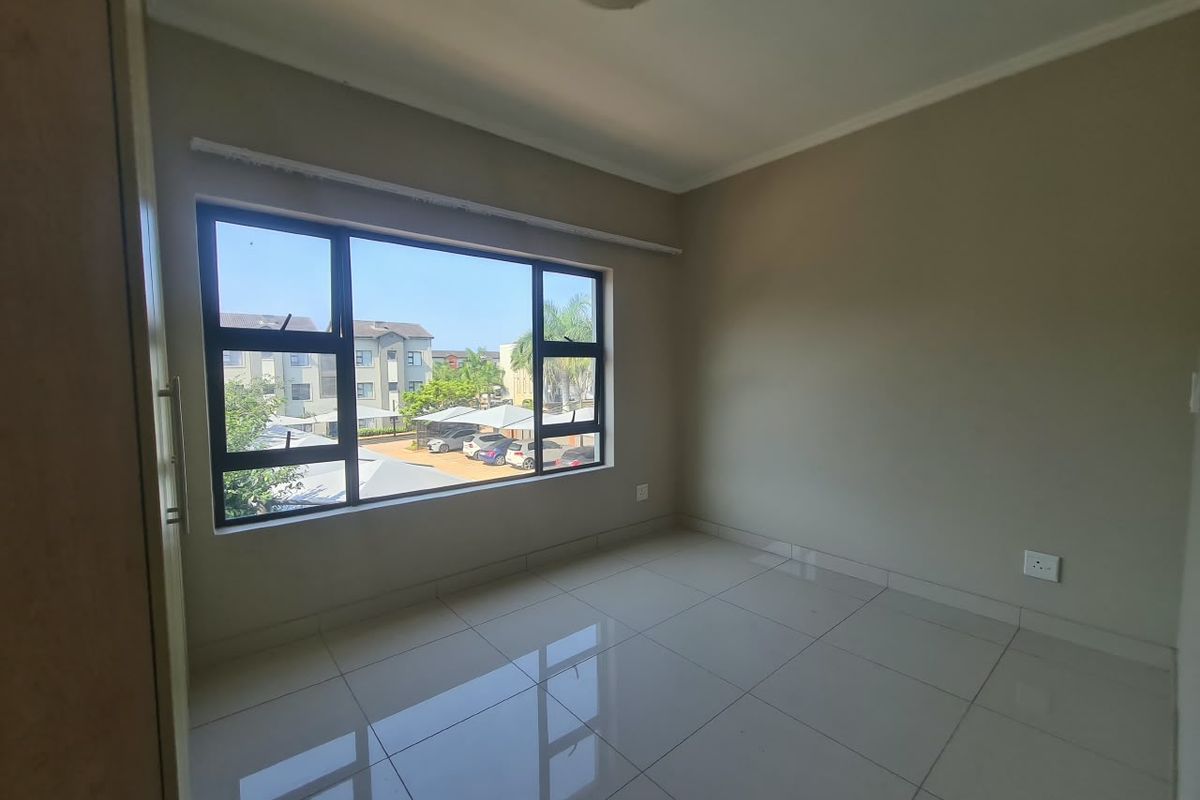 An empty bedroom featuring tiled flooring, neutral wall paint, and large aluminium-framed windows overlooking the complex parking area.