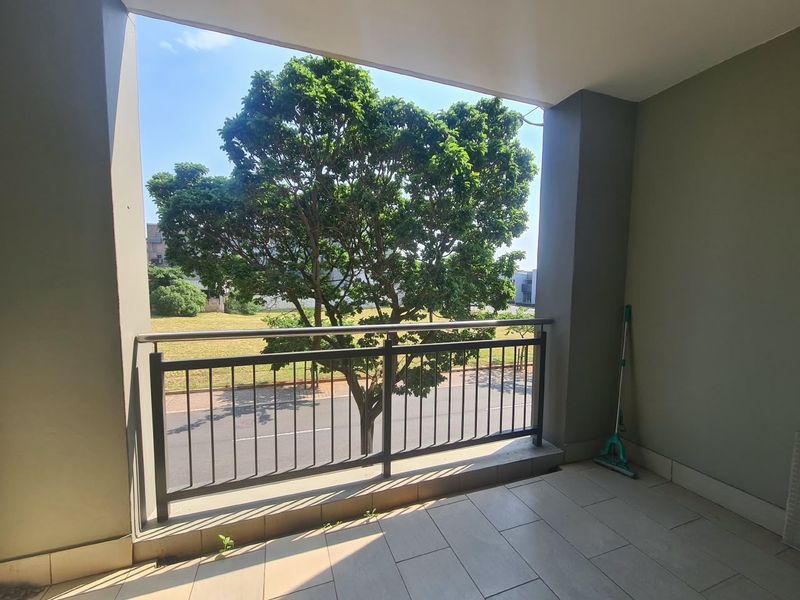A covered balcony featuring tiled flooring and a metal balustrade with a stainless steel handrail, offering an open view of the street and greenery.