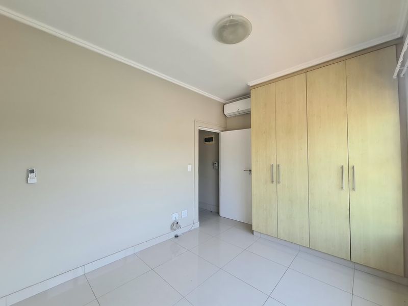 A bright, empty bedroom featuring light-toned built-in cupboards, tiled flooring, and a wall-mounted air conditioning unit.