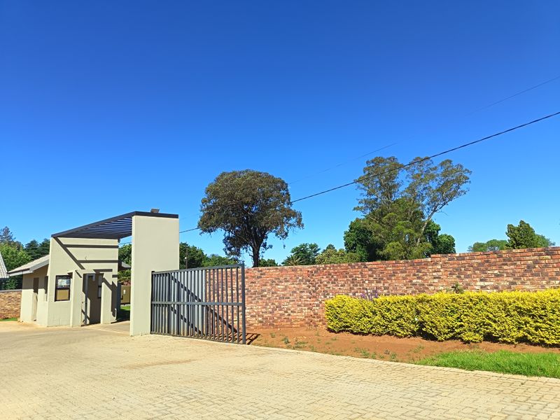 Front entrance of the townhouse complex featuring a paved driveway, brick boundary wall, metal security gate, and a modern guard house structure.