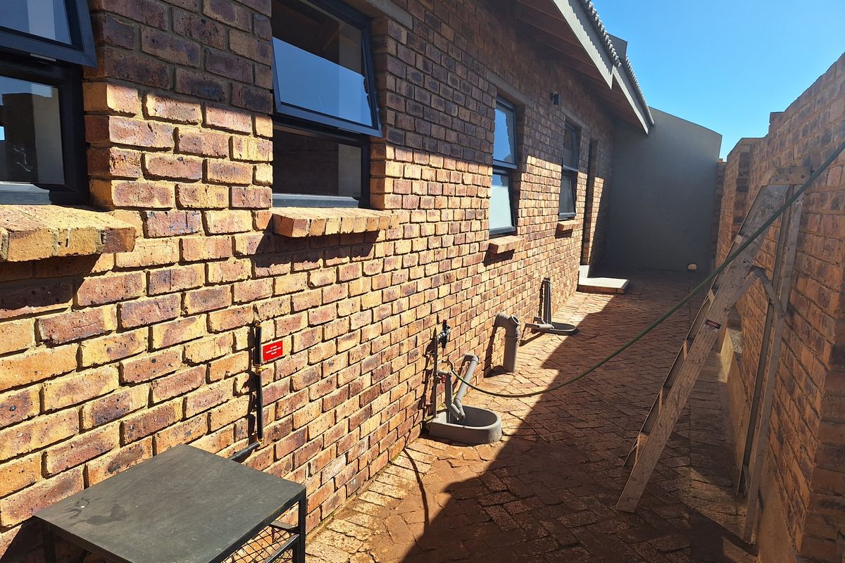 A side exterior view of the townhouse featuring face brick walls, aluminium window frames, and a paved walkway with exposed utility plumbing connections.