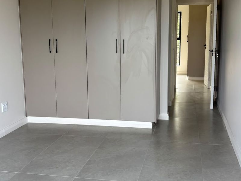A clean, empty bedroom featuring modern grey floor tiles and built-in cupboards, leading to a hallway and adjacent room.
