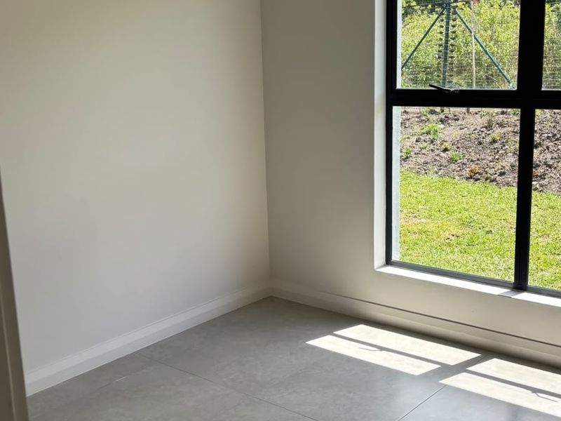 A bright, empty bedroom featuring modern large-format floor tiles and a large black aluminium window overlooking the garden.