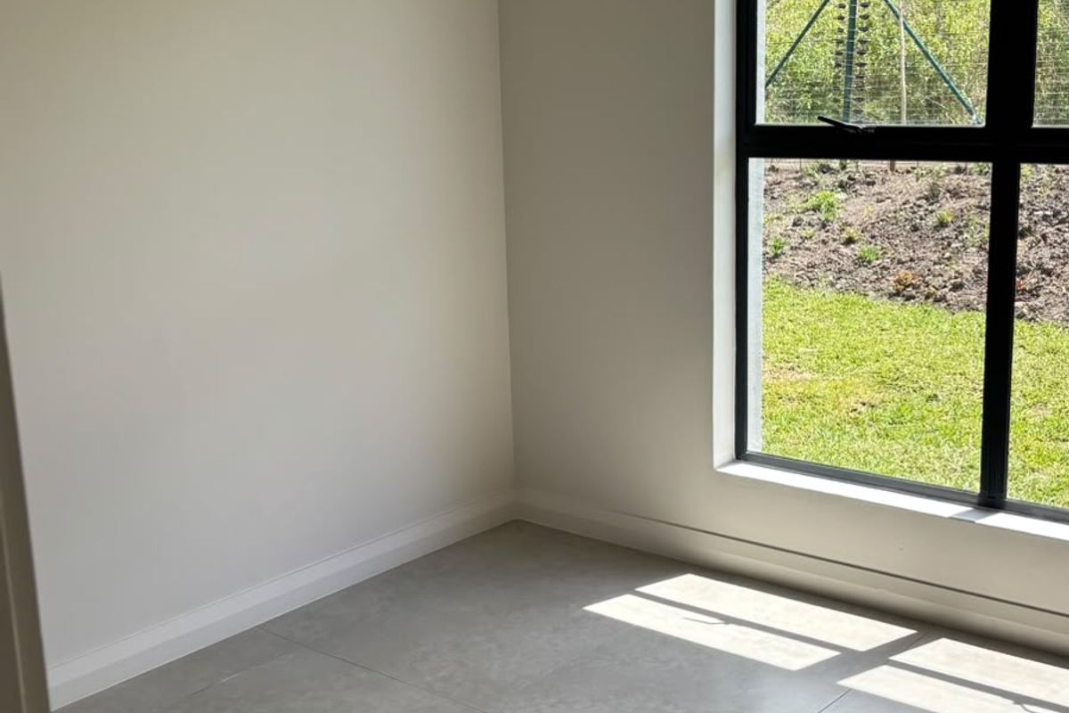 A bright, empty bedroom featuring modern large-format floor tiles and a large black aluminium window overlooking the garden.