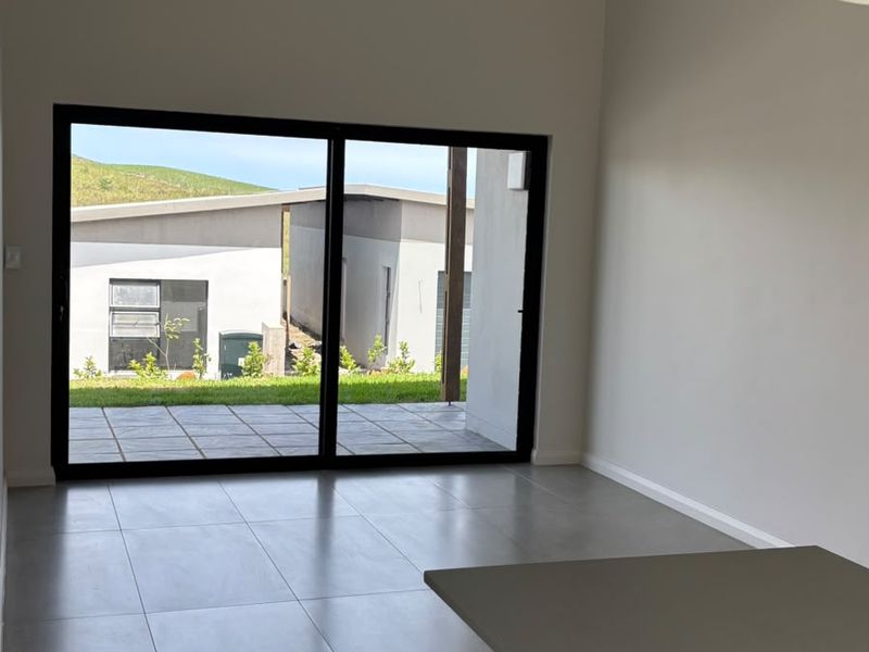 An open-plan living area featuring large sliding glass doors that open onto a paved patio, with neutral tiled flooring and recessed lighting.