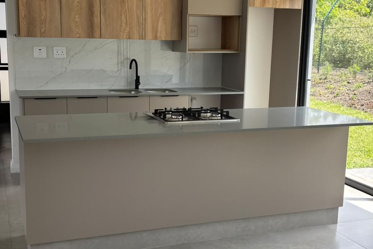 Modern kitchen featuring a central island with a gas hob, stone countertops, wood-grain cabinetry, and sleek pendant lighting, opening to an outdoor area.