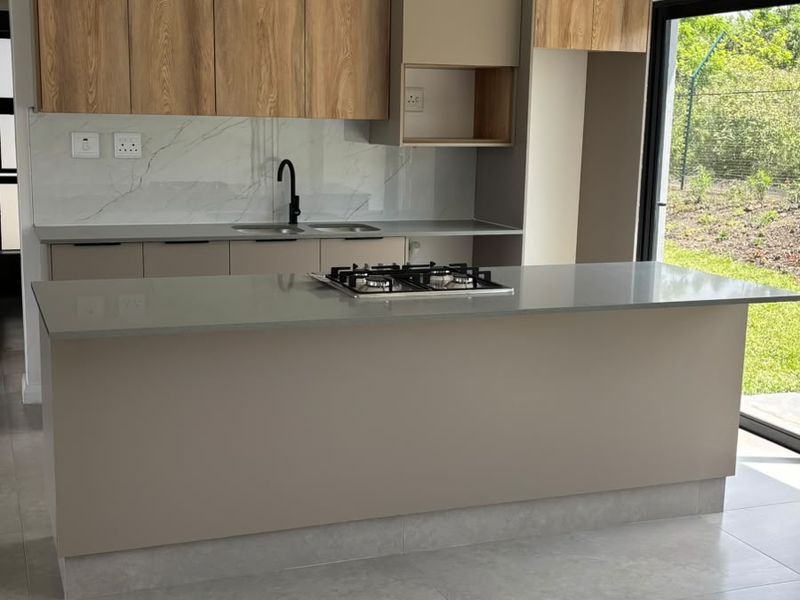 Modern kitchen featuring a central island with a gas hob, stone countertops, wood-grain cabinetry, and sleek pendant lighting, opening to an outdoor area.