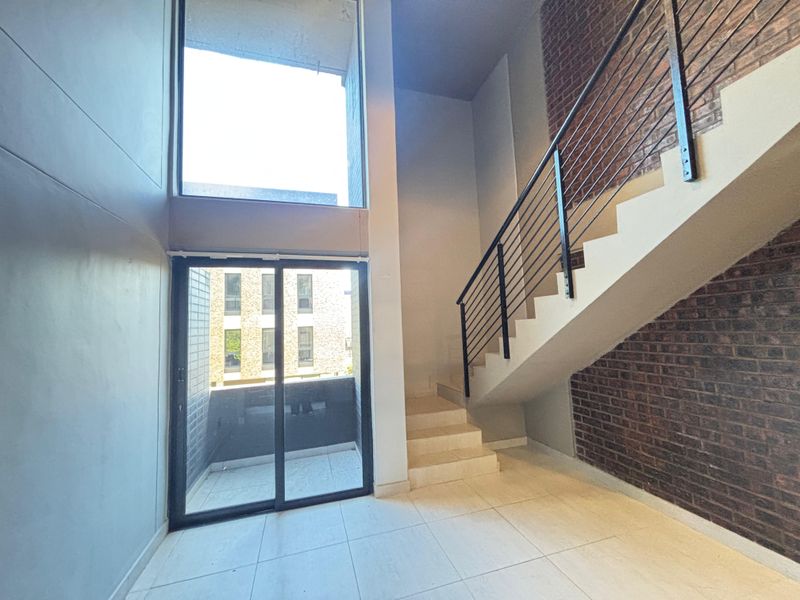 A bright, double-volume living area featuring modern tiled floors, an exposed brick feature wall, and a staircase with a sleek metal balustrade.
