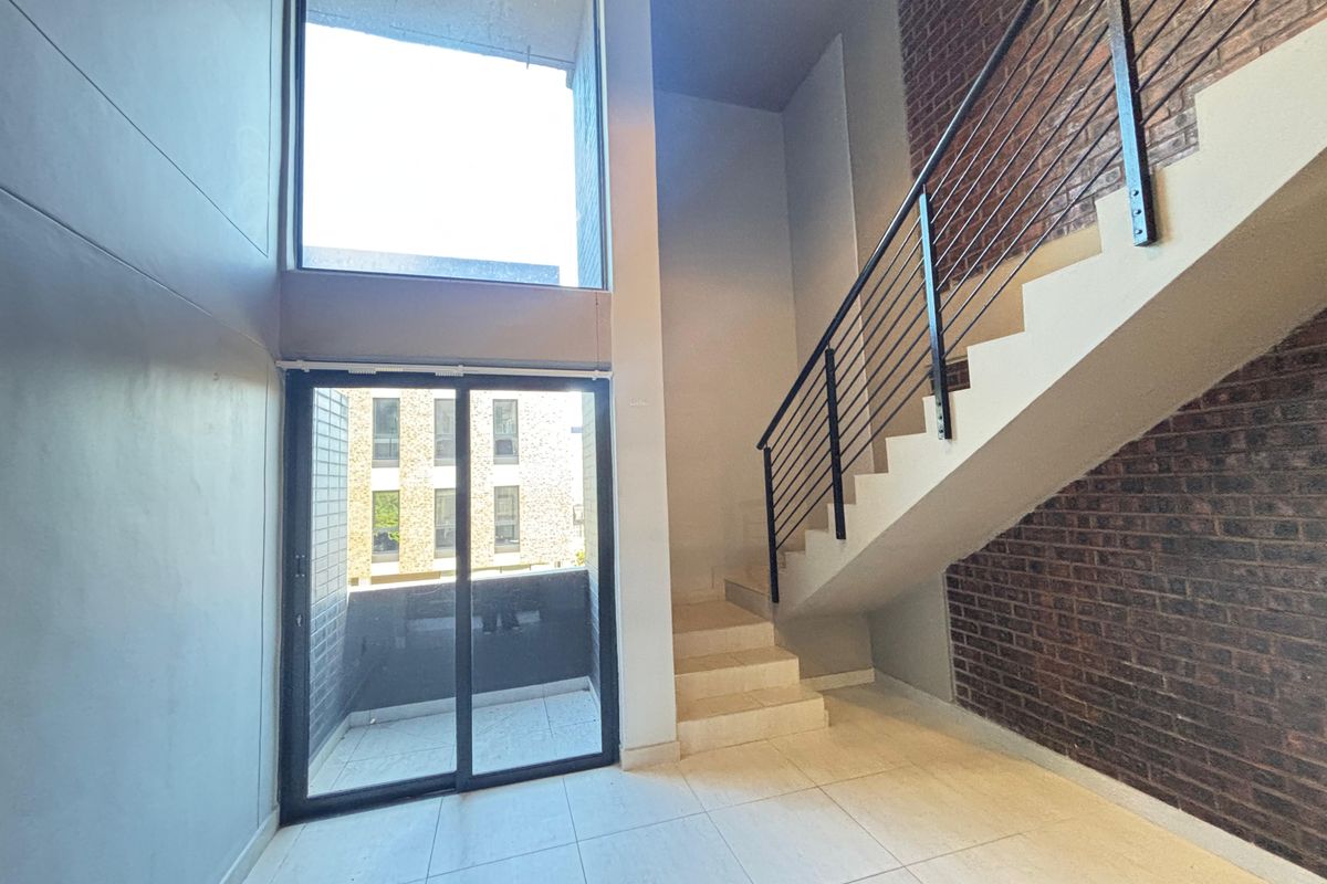 A bright, double-volume living area featuring modern tiled floors, an exposed brick feature wall, and a staircase with a sleek metal balustrade.