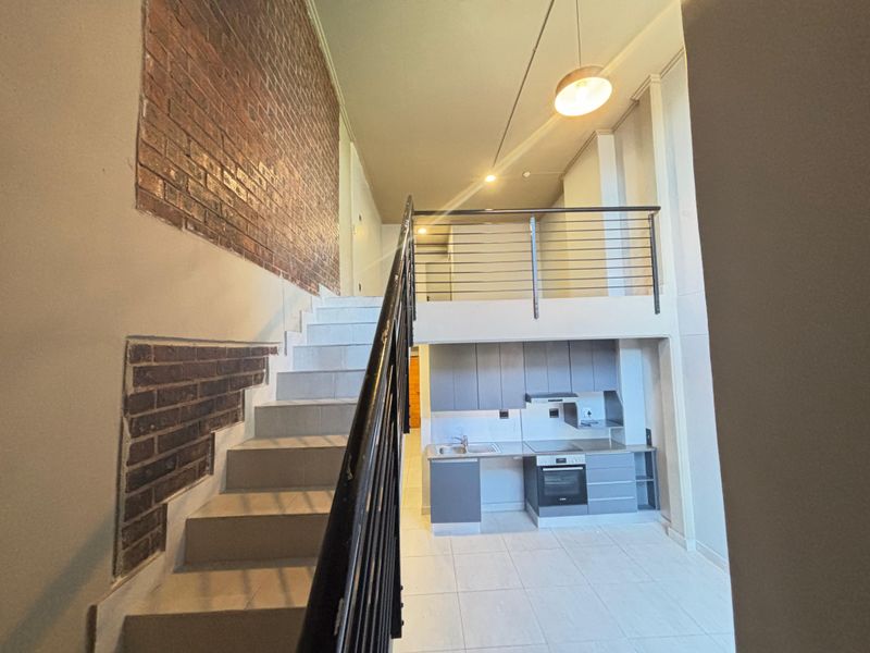 Modern open-plan apartment featuring a mezzanine level, exposed brick accents, a sleek kitchen with built-in appliances, and a staircase with metal balustrade.