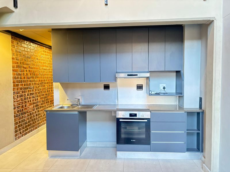 Modern kitchen featuring sleek grey cabinetry, stainless steel countertops, built-in oven, hob, and an industrial-style exposed brick feature wall.