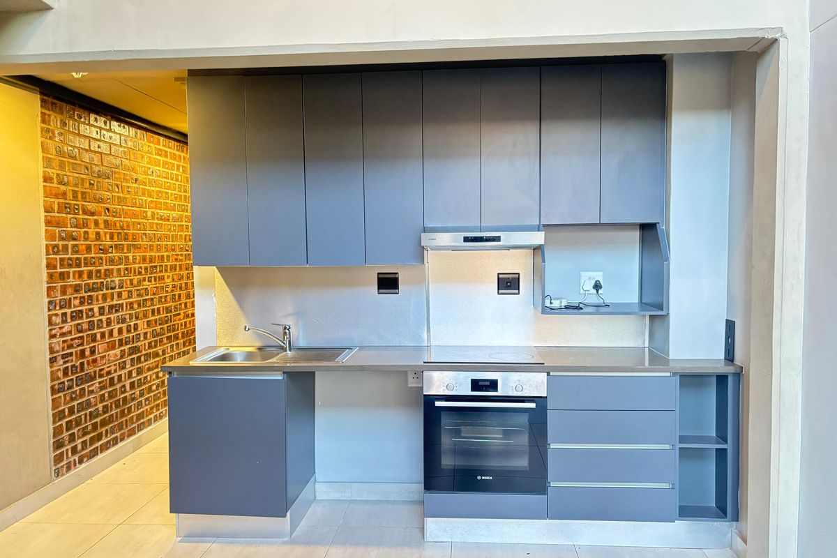 Modern kitchen featuring sleek grey cabinetry, stainless steel countertops, built-in oven, hob, and an industrial-style exposed brick feature wall.