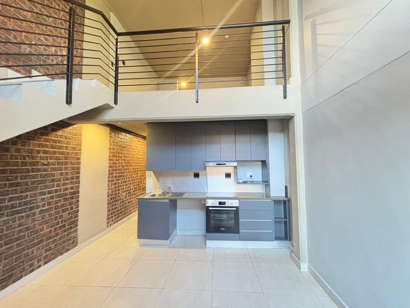 Open plan kitchen area featuring modern grey cabinetry, built-in oven, and extractor fan, set against an exposed brick wall with an overhead mezzanine level.