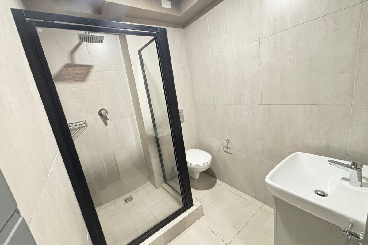 A modern, clean bathroom featuring a walk-in shower with a black-framed screen, a wall-mounted toilet, and a sleek vanity basin with neutral tiling.