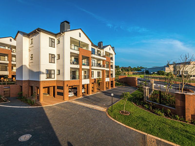 A modern multi-story apartment complex featuring a brick and plaster facade, private balconies, and a well-maintained paved driveway with covered parking areas.