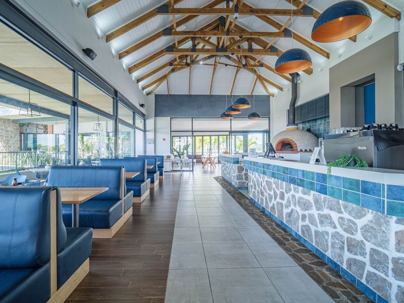 Spacious clubhouse interior featuring exposed timber trusses, a stone-clad service counter with a built-in pizza oven, and expansive glass doors.