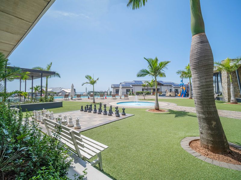A well-maintained outdoor recreational area featuring a swimming pool, artificial lawn, paved pathways, and a water feature, surrounded by landscaped palm trees.