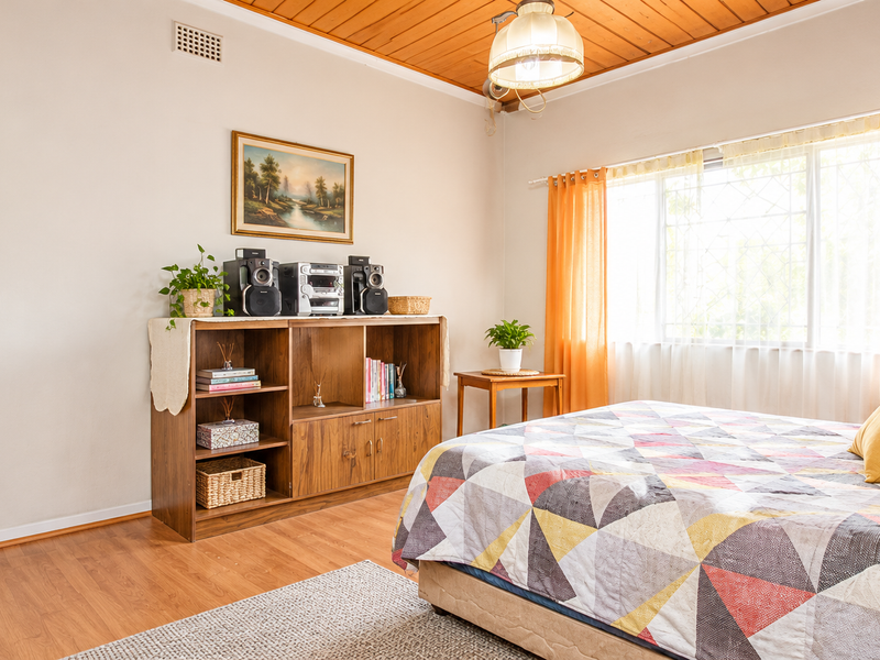 A well-lit bedroom featuring warm timber ceiling paneling, laminate flooring, and a large window with security bars, offering a comfortable and functional living space.