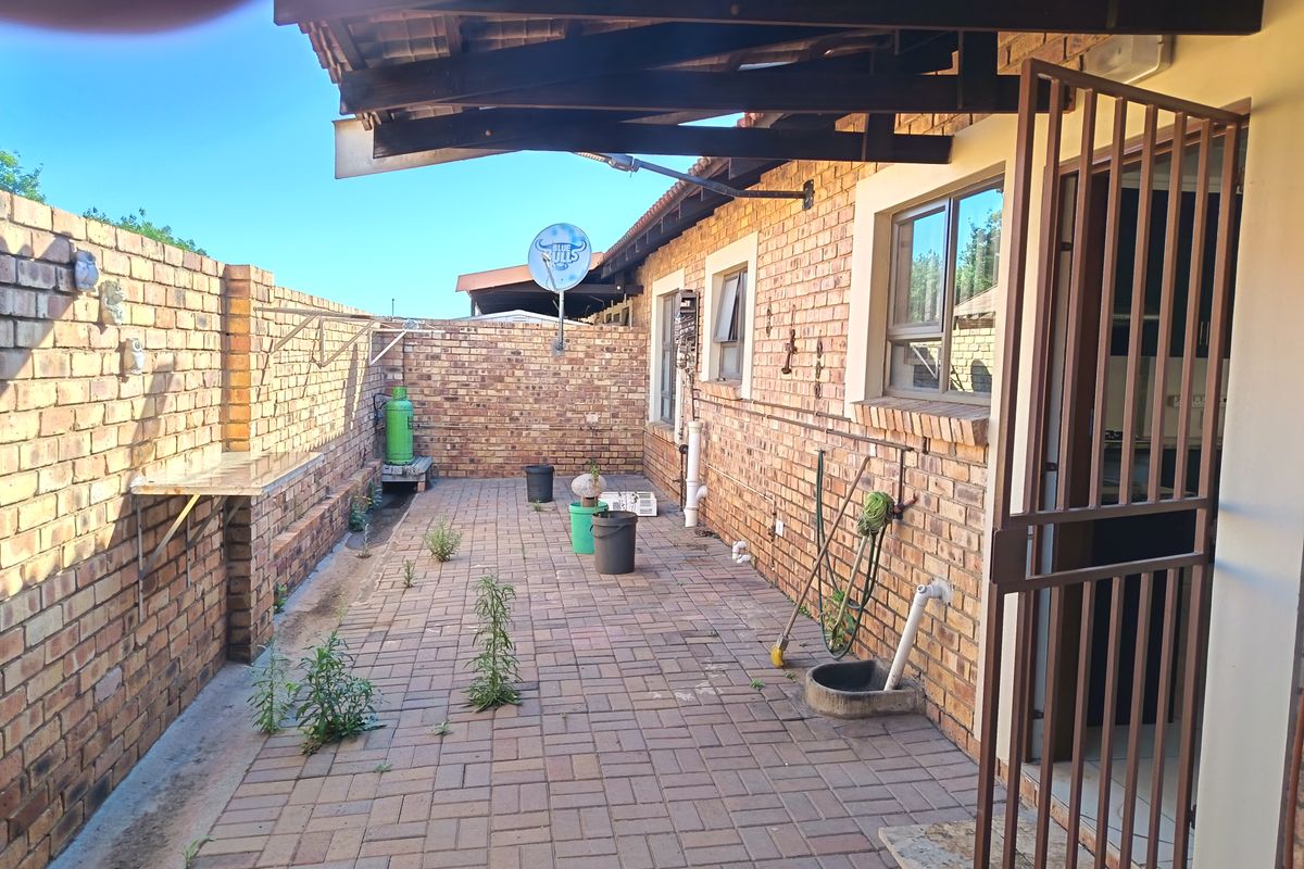A paved rear courtyard featuring a covered patio area, brick boundary walls, and direct access to the home via a security gate.