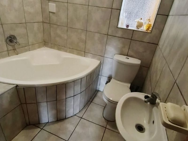 A compact bathroom featuring a corner bathtub, pedestal basin, and toilet, finished with neutral ceramic tiling throughout.