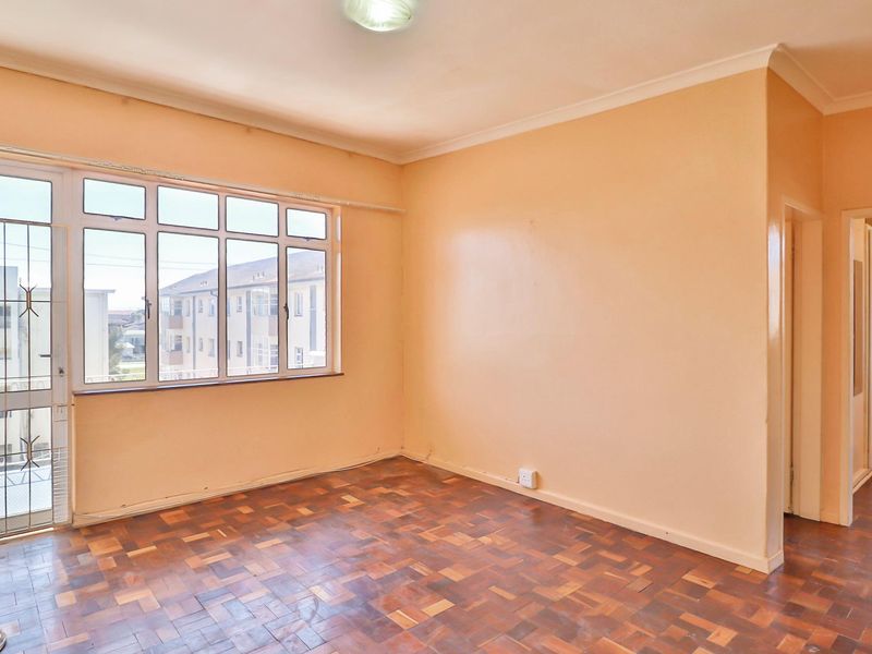 An empty living room featuring classic parquet flooring, a large window with a security gate, and neutral wall paint.