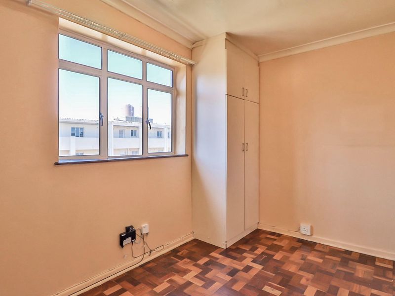 A bright, empty bedroom featuring classic parquet flooring, a built-in cupboard, and large aluminium-framed windows providing natural light.