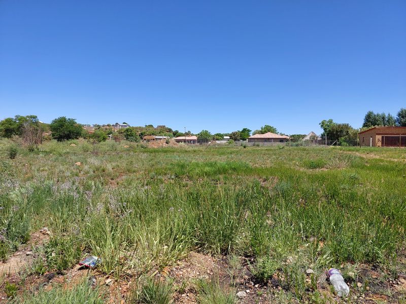 A vacant residential plot featuring natural grass coverage and perimeter wire fencing, situated in a developed neighborhood setting.