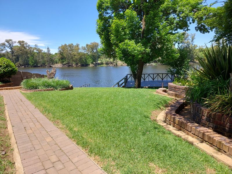A scenic view of the riverfront garden featuring a brick paved pathway, tiered brick retaining walls, and a private wooden jetty overlooking the water.