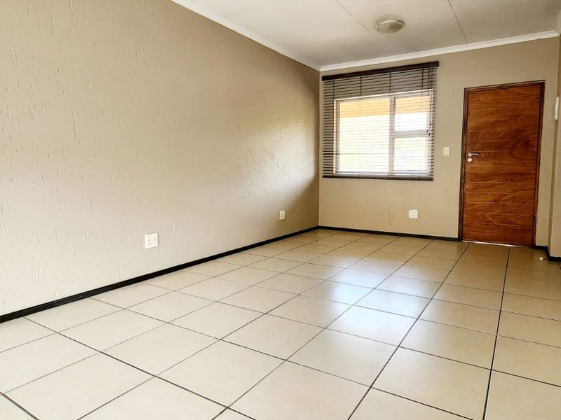 A bright, empty living room featuring neutral wall tones, tiled flooring, and a wooden door with natural light from the window.