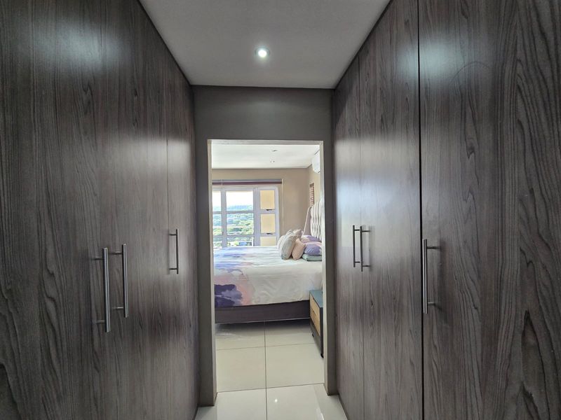 A bedroom view through a dressing area featuring extensive built-in cupboards with modern wood-grain finish and sleek handles, leading into a bright sleeping space.