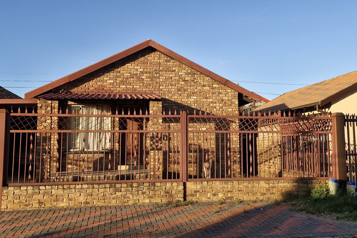 Front elevation of a face brick house featuring a secure metal perimeter fence, gated entrance, and paved driveway area.