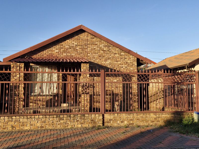 Front elevation of a face brick house featuring a secure metal perimeter fence, gated entrance, and paved driveway area.