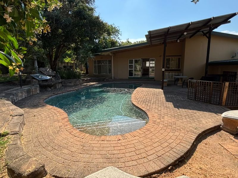 Outdoor pool area featuring a curved swimming pool, brick paving, and a covered patio space, set within a landscaped garden environment.