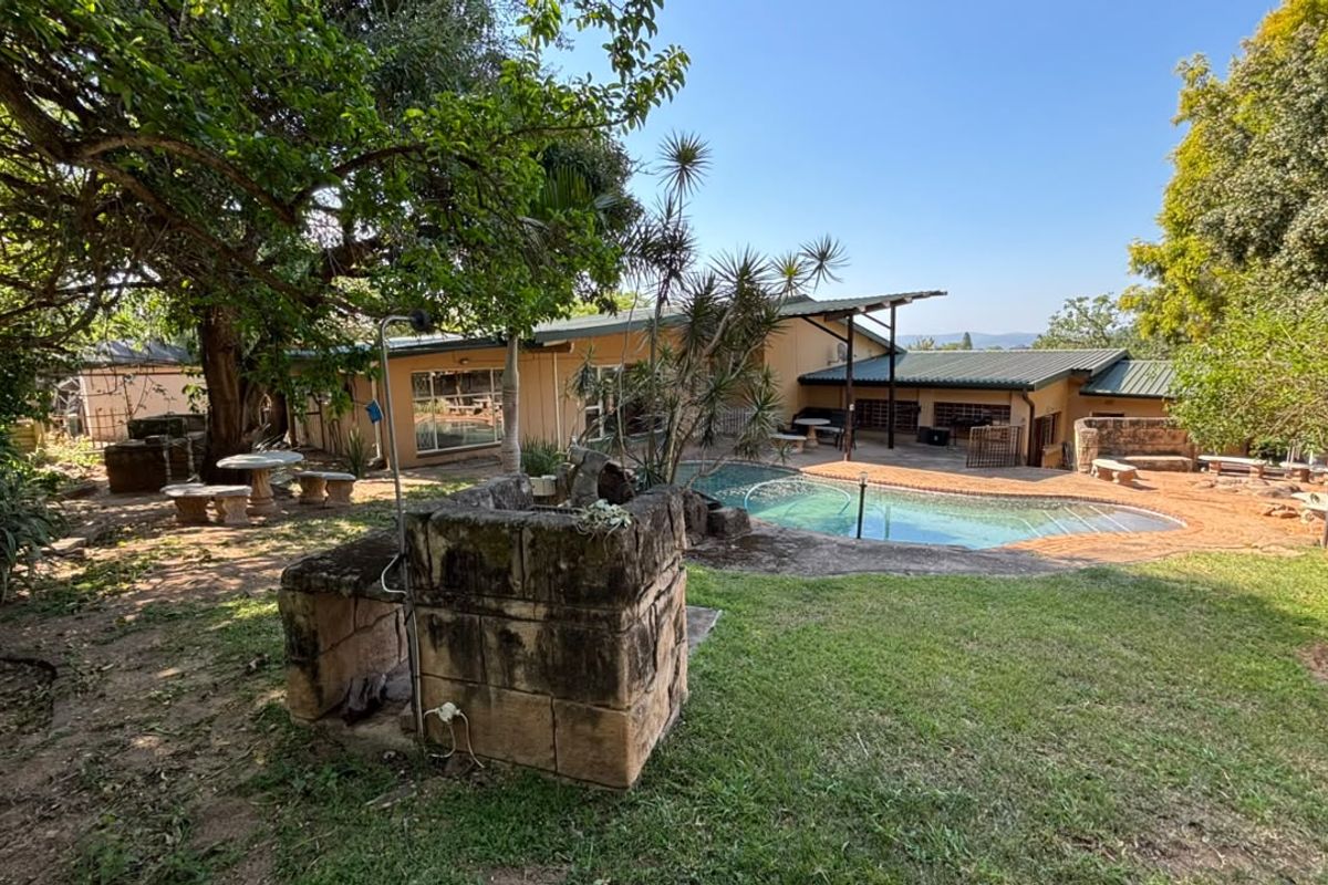 Rear garden view featuring a swimming pool, covered patio area, and a stone built-in braai, set within a mature landscaped yard.