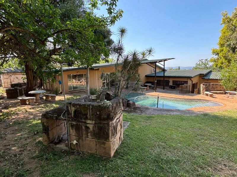 Rear garden view featuring a swimming pool, covered patio area, and a stone built-in braai, set within a mature landscaped yard.