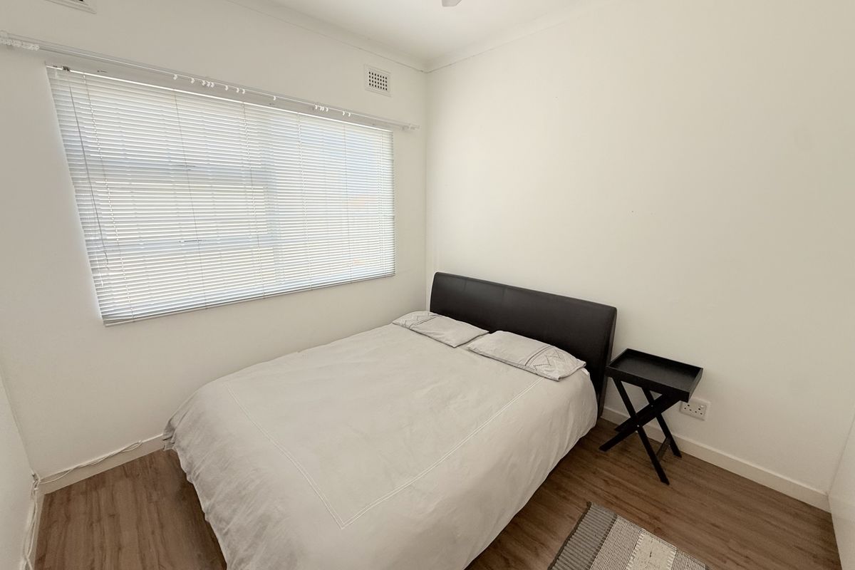 A clean and simple bedroom featuring laminate flooring, a ceiling fan, and large windows with venetian blinds for natural light.