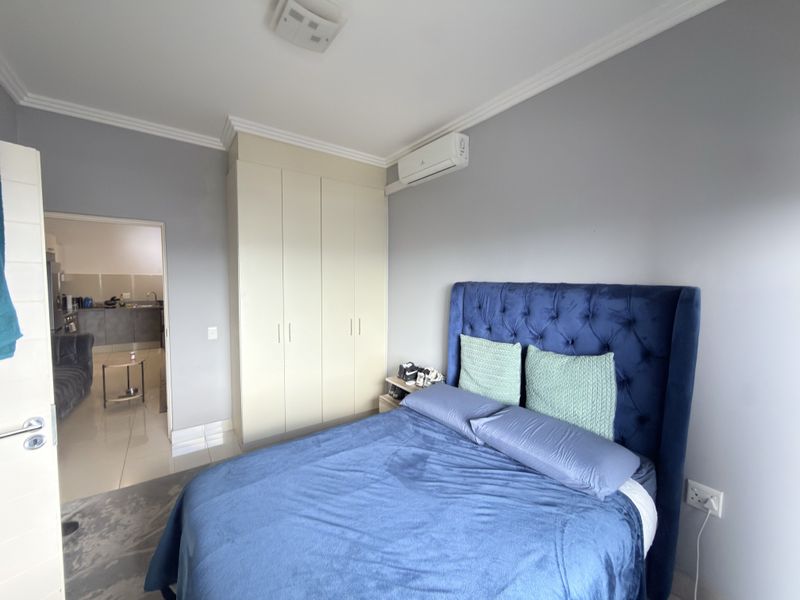 A well-maintained bedroom featuring built-in wardrobes, a split-system air conditioner, and neutral wall finishes, with an adjacent view into the kitchen area.