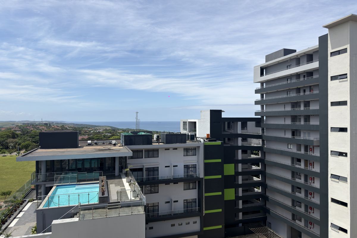 An elevated aerial view of a modern apartment complex featuring a rooftop swimming pool, glass balustrades, and surrounding coastal landscape.
